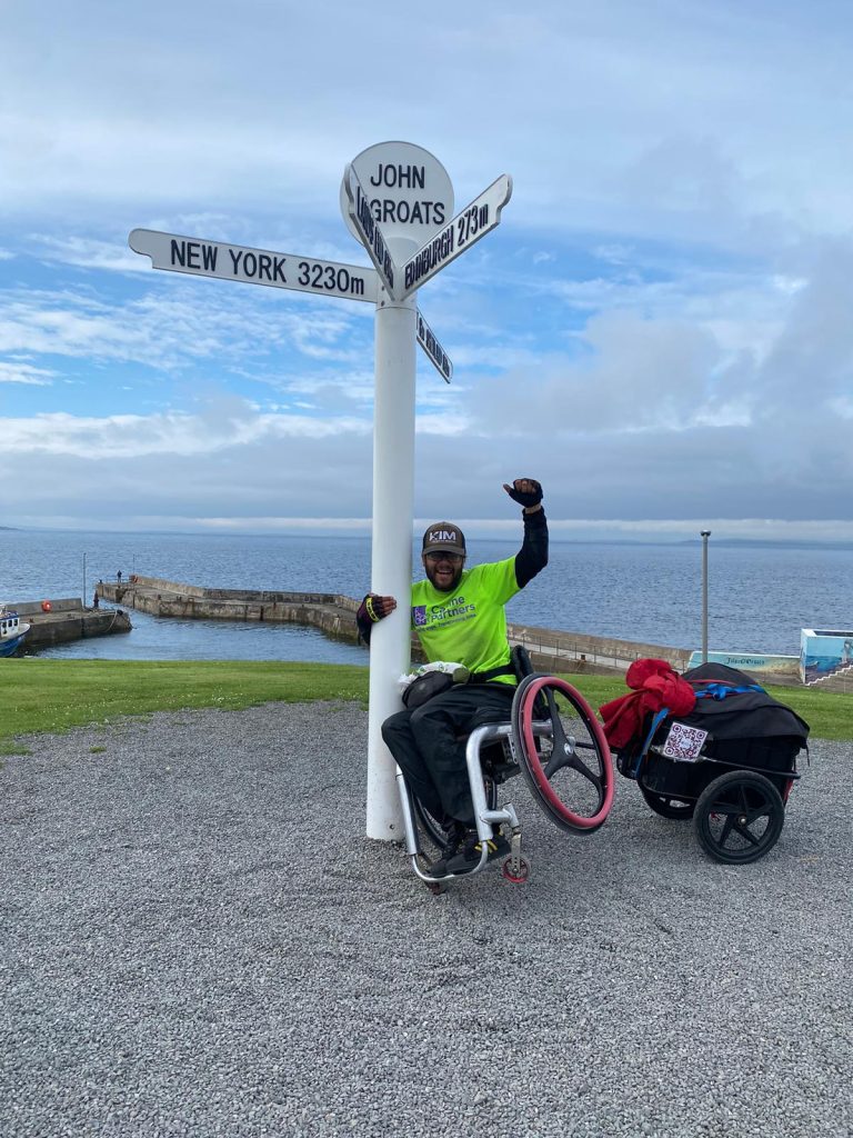 A man in a wheelchair holding on to a sign on top of a poll. The sign reads JOHN O'GROATS and has distance markers pointing the way to various points on the globe.
