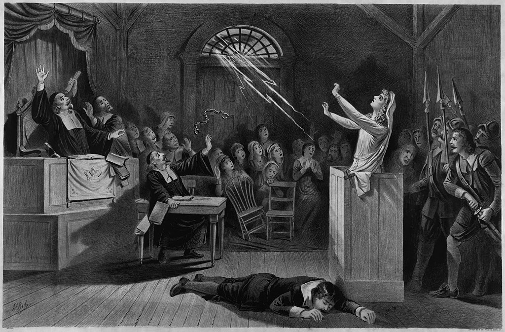 A women stands in the dock of a court with a judge looking on as rays of light point towards her from a window.